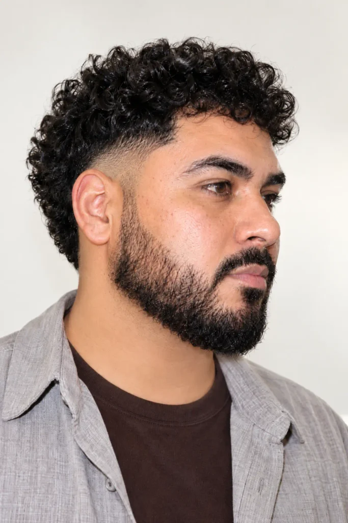 Man with a textured curly top, mid burst fade, and full beard blended neatly into the sideburns for a balanced men's haircut.